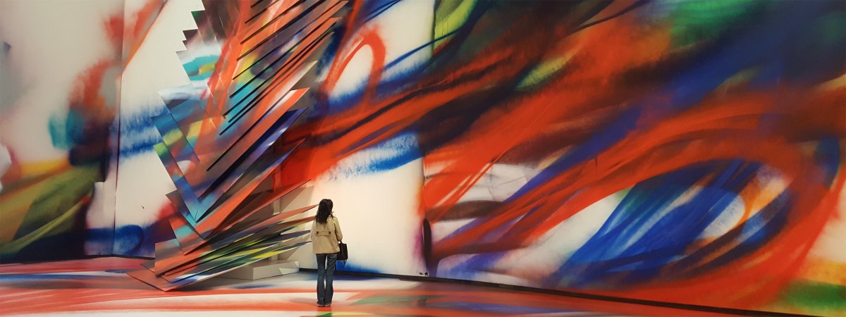 A woman stands in a large gallery space in front of a massive abstract wall mural with sweeping brushstrokes in red, blue, orange, green, and white.