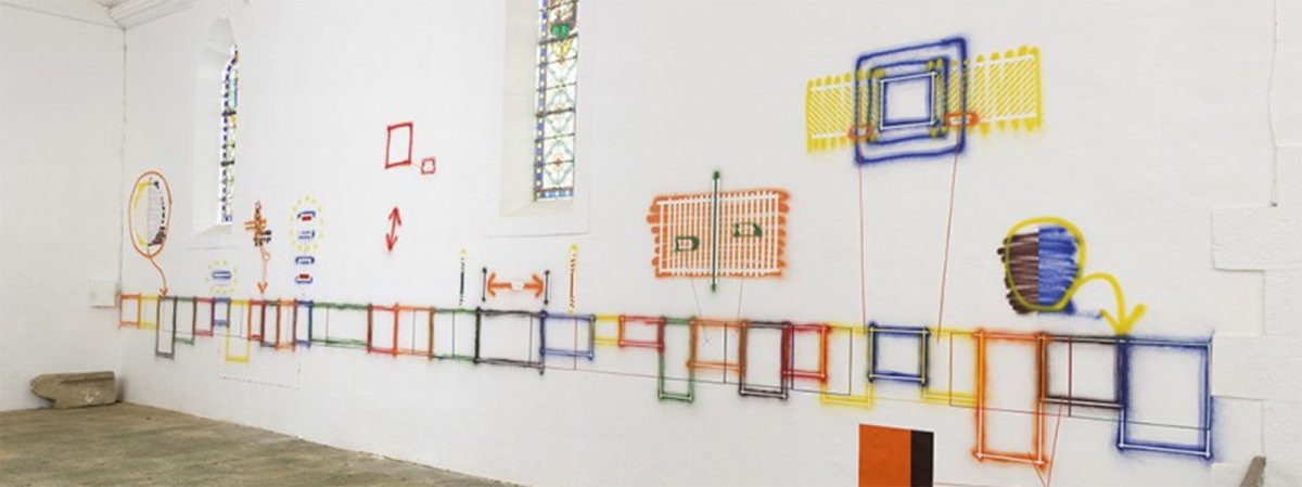 A long, colorful abstract wall installation made of many connected rectangular and looped elements in red, yellow, blue, green, and orange, mounted on a white gallery wall in a gallery space.